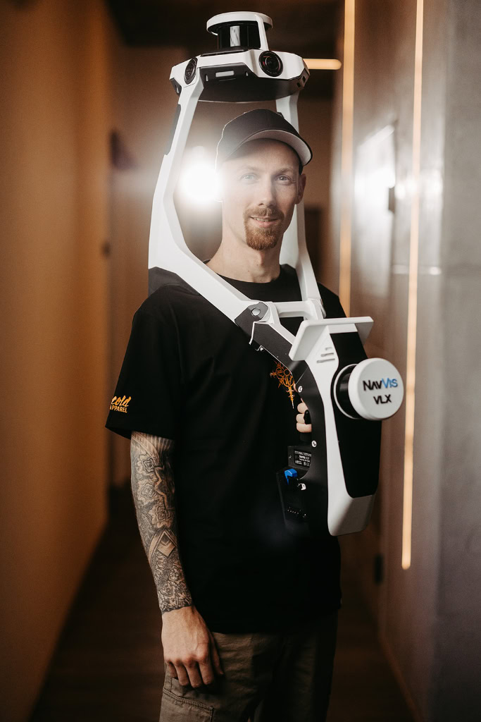 Ein Mann mit Tätowierungen steht in einem Flur mit einem 3D-Laserscanner auf dem Kopf.