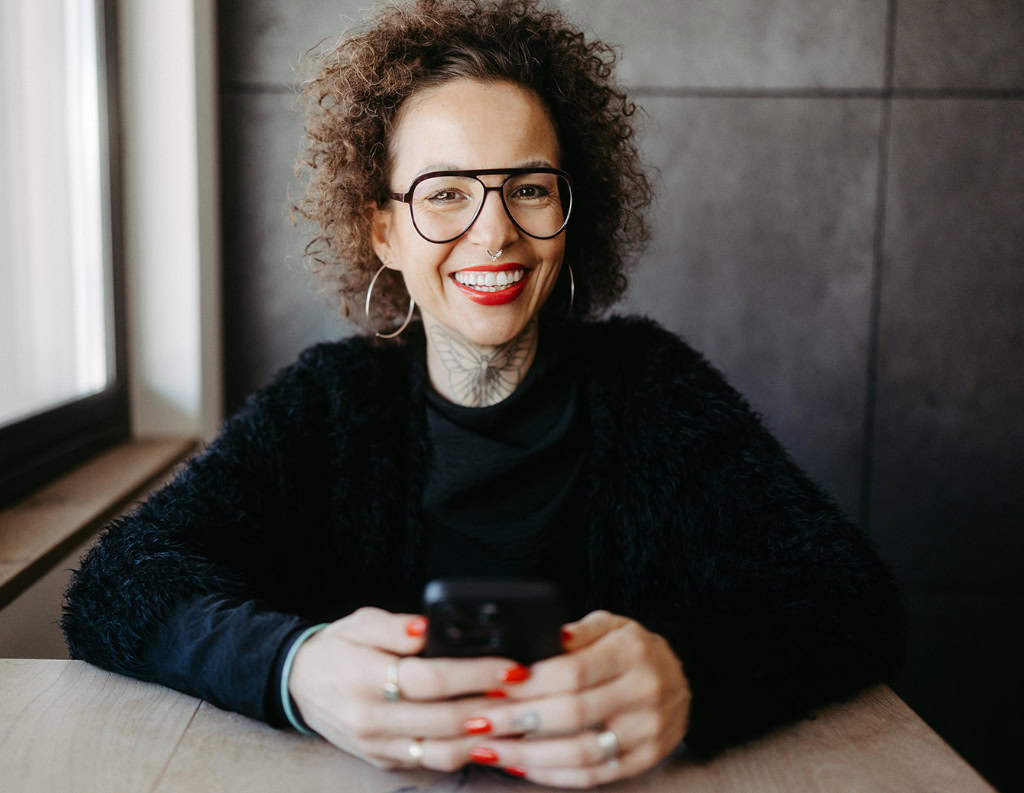 Eine Frau mit lockigem Haar und Brille sitzt mit einem Smartphone an einem Tisch.
