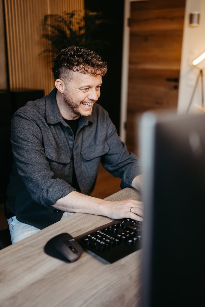 Ein Mann lächelt, während er an einem Schreibtisch vor einem Computer sitzt.